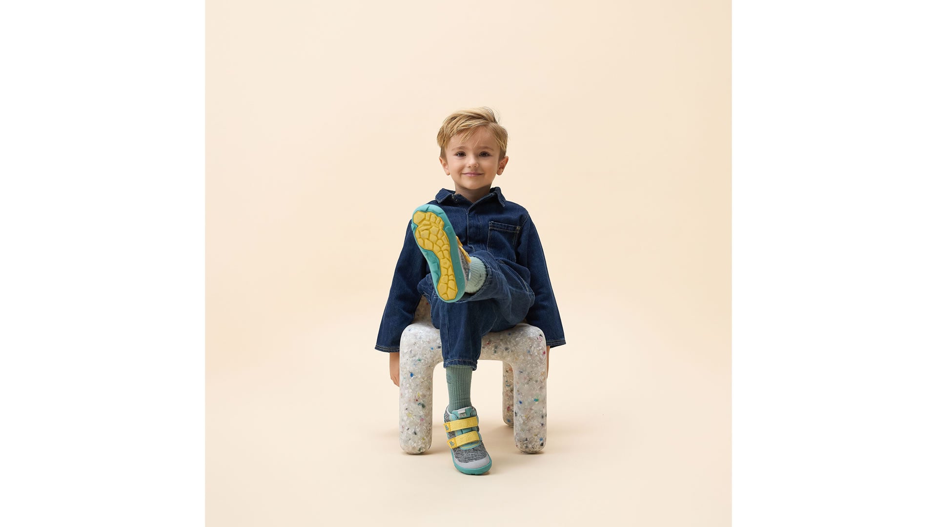 Affenzahn Low Boot Knit Happy - Rhino šedo-zelené zapínáním na suchý zip ve žluté barvě chlapec sedící na židličce se zvednutou levou nohou je vidět detail podrážky levé nohy a nárt pravé nohy